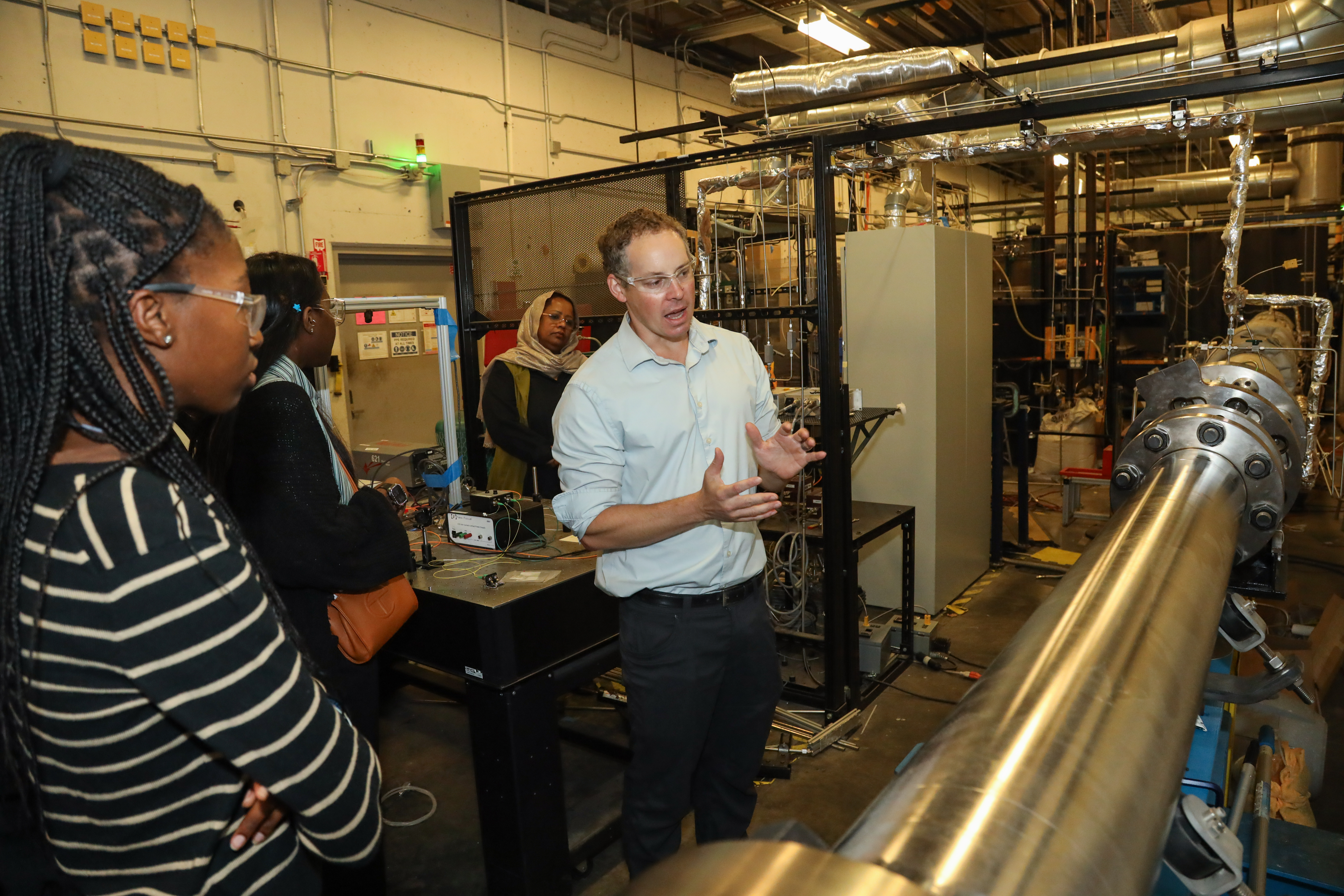 People wearing safety glasses listen as a researcher gestures toward a large metal apparatus in an industrial lab with pipes and equipment.