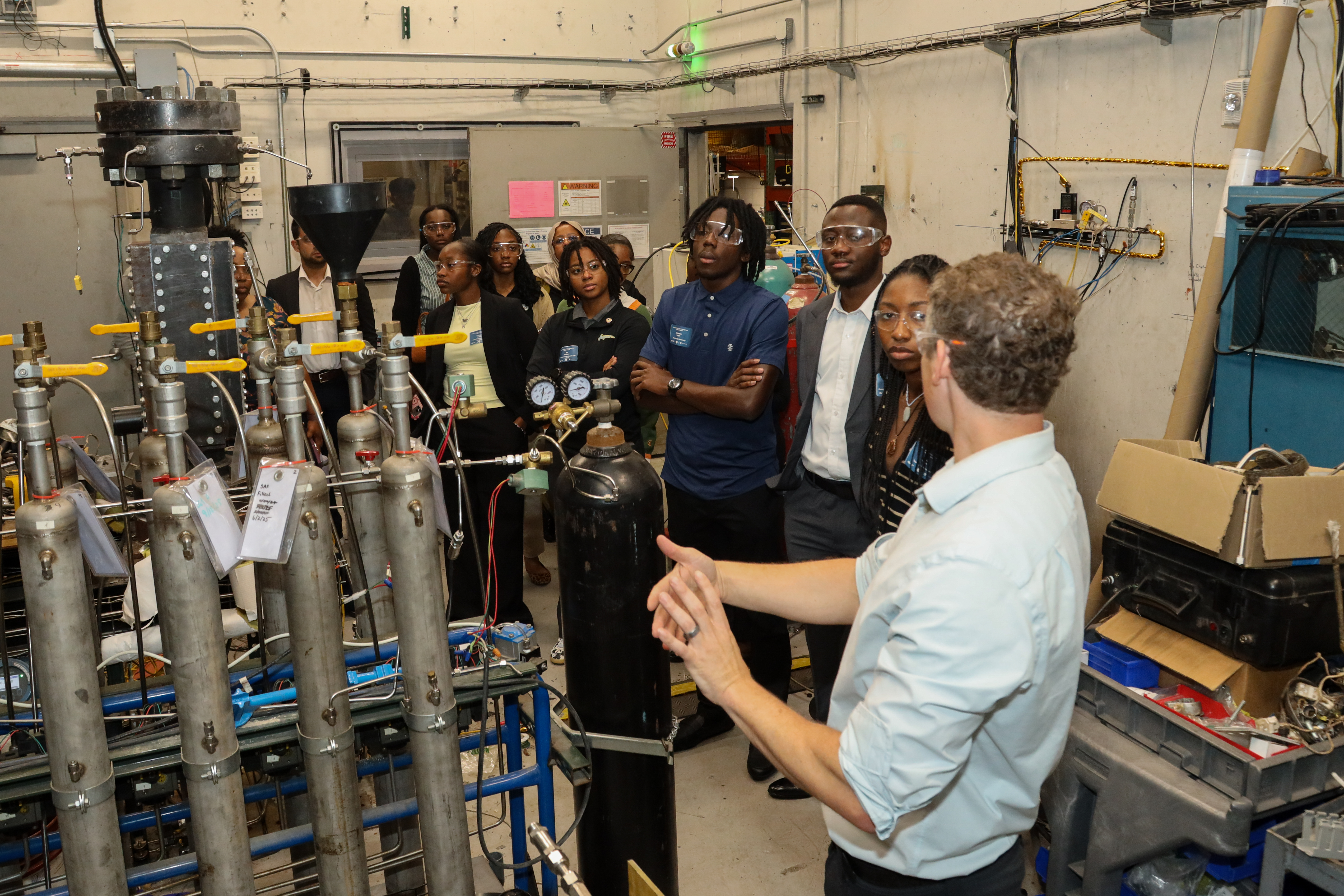 A group in safety glasses gathers around gas cylinders and instruments while a researcher explains a laboratory setup.