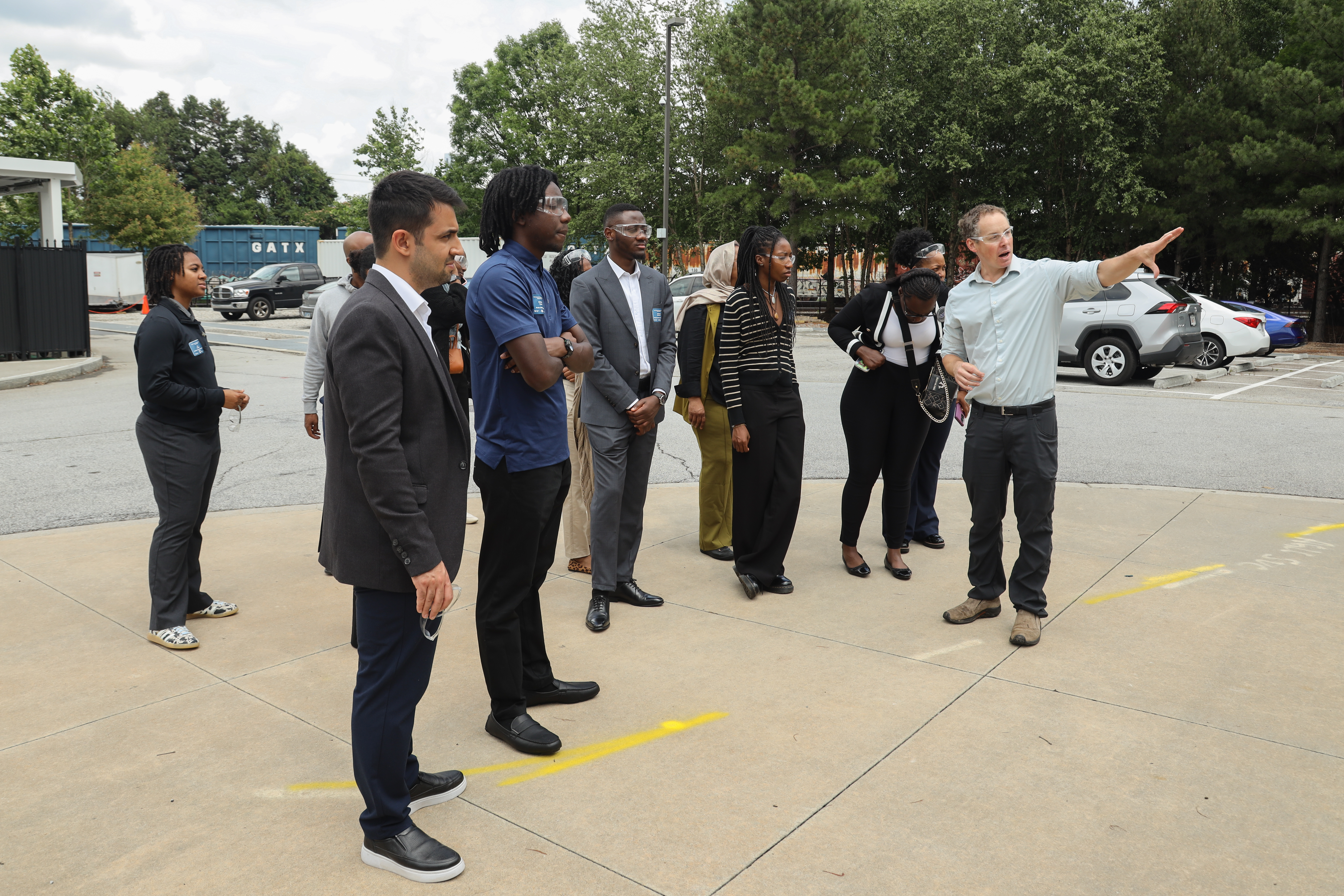 A group stands outside as a researcher gestures and explains something near a building, with parked cars and trees in the background.